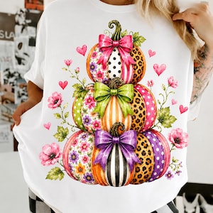 May include: A white t-shirt featuring a graphic of three stacked pumpkins. The pumpkins are decorated with various patterns including leopard print, polka dots, stripes, and floral designs, each adorned with a large pink, green, or purple bow.