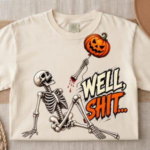 May include: A cream-colored t-shirt featuring a graphic of a skeleton sitting on the ground. The skeleton holds a string attached to an orange jack-o'-lantern balloon. The text "WELL, SHIT..." is printed in large, bold, orange and black letters.