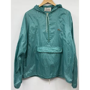 Könnte beinhalten: Eine türkisfarbene Kapuzen-Windjacke zum Überziehen mit halbem Reißverschluss, weißen Kordelzügen, einer großen Fronttasche mit Klappe und einem kleinen gestickten Krokodil-Logo auf der Brust. Das Innenetikett zeigt den Text ZOD LACOSTE.