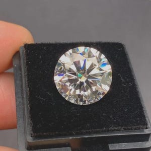 May include: A round, brilliant-cut, colourless gemstone with multiple facets that reflect light, creating a sparkling effect. The stone is securely set within a square, black velvet-lined display box, which provides a high-contrast backdrop.