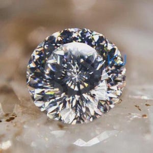 Può includere: Una pietra preziosa rotonda a taglio brillante con un aspetto chiaro e incolore e numerose sfaccettature triangolari che creano un alto livello di riflessione della luce e brillantezza. La pietra presenta un motivo geometrico nitido.