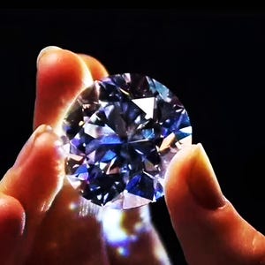 Può includere: Una pietra preziosa rotonda a taglio brillante con un aspetto chiaro e traslucido. La pietra presenta numerose sfaccettature triangolari e a forma di aquilone che riflettono la luce, creando un effetto scintillante con riflessi blu e bianchi su sfondo scuro.