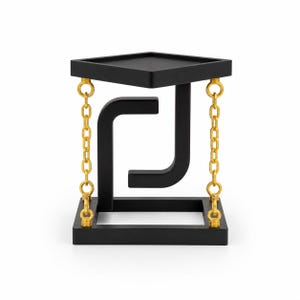 May include: A black square-shaped table with a matte finish, featuring a central J-shaped support structure. The top platform is suspended above the base by four gold-colored metal chains attached to the corners, creating a floating visual effect.