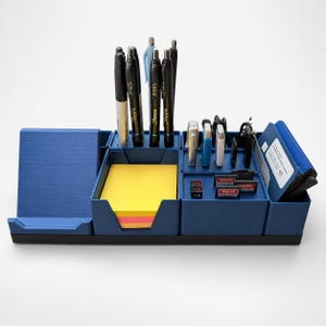 May include: A blue desk organizer featuring a dedicated slot for a smartphone, a central compartment filled with yellow sticky notes, and various sections holding pens, USB drives, SD cards, and a small blue wallet on the far right side.