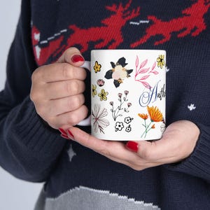May include: A white ceramic mug featuring a colorful botanical pattern with various illustrated flowers in shades of yellow, orange, pink, and black. The design includes delicate stems and leaves, along with the partial cursive text Antho in blue.