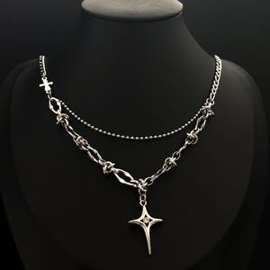 Op de afbeelding: Een zilverkleurige gelaagde ketting met een fijne bolletjesketting en een dikkere, getextureerde ketting met knoopachtige schakels. Een kleine kruisbedel is aan de zijkant bevestigd en er hangt een vierpuntige, stervormige kruishanger met een steen.