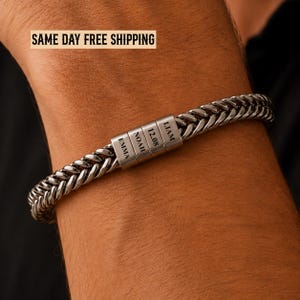 May include: A silver-toned metal wheat chain bracelet featuring four rectangular beads engraved with the names EMMA, NOAH, the date 12.08, and the name LIAM. The bracelet has a polished finish and a secure, integrated clasp mechanism.