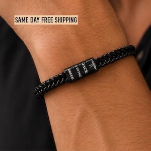 May include: A black braided bracelet featuring three rectangular metal beads. The beads are engraved with the text "JACK A DYING BREED" and a small lightning bolt icon. The bracelet has a woven texture and a polished black finish.