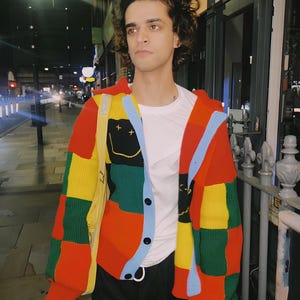 May include: A long-sleeve button-up cardigan featuring a vibrant patchwork design of red, yellow, green, and light blue blocks. The knit garment includes a black panel with a yellow embroidered smiley face and a yellow strap accessory.