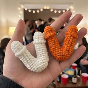 May include: Two small, V-shaped crochet items, one in white and one in orange. Each piece features a textured, yarn-stitched surface with rounded ends and a distinct ridge detail near the top of each arm, held in an open palm.
