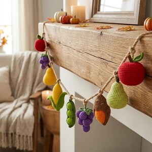 Peut inclure: Une guirlande décorative composée de divers fruits et légumes crochetés, incluant une pomme rouge, du raisin violet, une poire jaune, un épi de maïs jaune, des petits pois, un gland marron et une poire vert clair, suspendus sur une corde en jute.