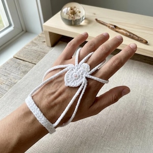May include: A white crocheted hand ornament featuring a central spider motif with eight legs extending to a wrist band and finger loops. The intricate thread work forms a delicate web-like structure that covers the back of the hand.