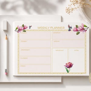 Pode incluir: Um bloco de notas semanal horizontal tamanho A5 com uma borda geométrica dourada, ilustrações em aquarela de flores de magnólia rosa e beija-flores. O layout inclui seções para cada dia da semana, além de caixas para notas importantes.