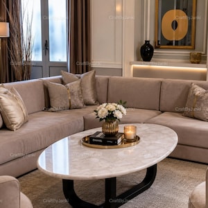 May include: An oval coffee table with a white stone top and a black base, featuring a gold tray holding a Chanel book, a glass candle, and a vase of white flowers. The table is positioned in front of a large beige sectional sofa with decorative pillows.