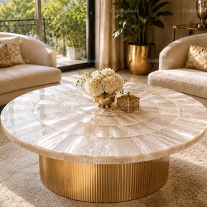May include: A round coffee table featuring a white, iridescent, shell-tiled tabletop arranged in a radial pattern, supported by a cylindrical, vertical-ribbed gold metal base. The surface holds a small gold bowl with white flowers and a gold box.