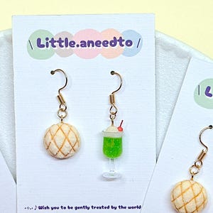 May include: A pair of gold-toned hook earrings featuring a miniature beige melon pan and a green cream soda float with a red cherry on top. The earrings are displayed on a white card labeled Little.aneedto with the text Wish you to be gently treated by the world.