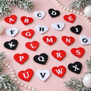 Puede incluir: Una colección de parches bordados en forma de corazón con las letras del alfabeto en blanco o negro. Los parches están dispuestos de forma dispersa, y cada corazón muestra una letra mayúscula individual en un color contrastante.
