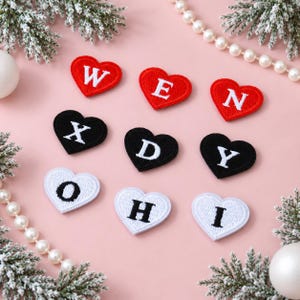 May include: A collection of nine heart-shaped embroidered patches arranged in three rows. The top row features red hearts with white letters W, E, and N. The middle row has black hearts with white letters X, D, and Y. The bottom row has white hearts with black letters O, H, and I.