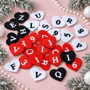 Puede incluir: Una colección de parches bordados en forma de corazón en negro, blanco y rojo. Cada parche presenta una letra o número bordado en un color contrastante. Los parches están esparcidos sobre una superficie, mostrando varios caracteres, incluyendo letras como E, Z, U, L, S, O, R, J, I, C, H, F, V, Q y números como 9, 6, 5, 8, 3.