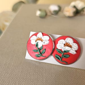 May include: A pair of round stud earrings featuring a vibrant red background with a raised white flower design. Each flower has a textured yellow centre and green stems with leaves. The earrings are set in a thin silver-toned metal frame.
