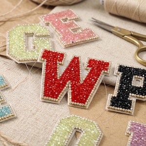May include: A collection of varsity-style letter patches featuring sparkling rhinestones in various colors including red, black, light green, and pink. Each letter is outlined with a border of small clear crystals, creating a textured, reflective surface.