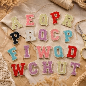 May include: A collection of varsity-style alphabet patches featuring a textured, glittery surface and a gold-toned beaded border. The letters appear in various colours including red, yellow, pink, blue, purple, black, and silver, arranged in rows.