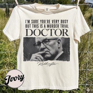 May include: An ivory short-sleeve t-shirt featuring a black and white photographic print of a man wearing glasses. The text reads: I'M SURE YOU'RE VERY BUSY BUT THIS IS A MURDER TRIAL DOCTOR and the name Reddington in a cursive script below.