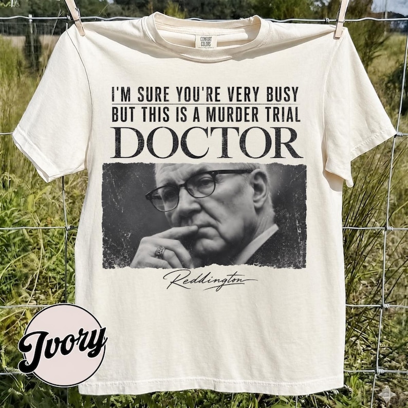 May include: An ivory short-sleeve t-shirt featuring a black and white photographic print of a man wearing glasses. The text reads: I'M SURE YOU'RE VERY BUSY BUT THIS IS A MURDER TRIAL DOCTOR and the name Reddington in a cursive script below.