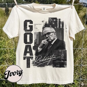 May include: An ivory short-sleeve t-shirt featuring a black and white graphic print of a man in a suit and glasses. The text reads "GOAT", "GREATEST OF ALL TIME", and "Reddington" in a stylized script font. The design includes a city building.