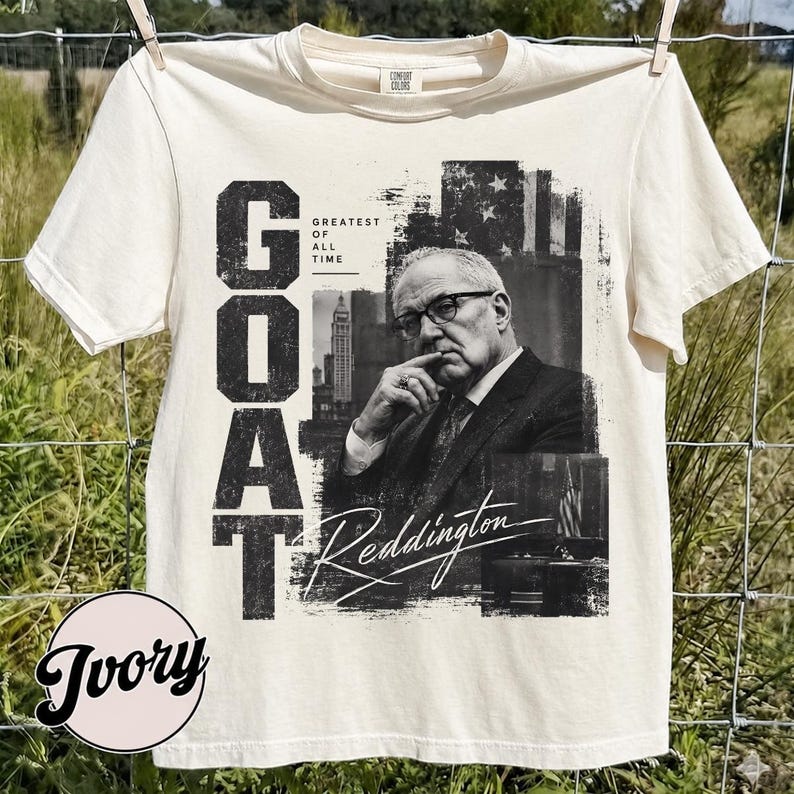 May include: An ivory short-sleeve t-shirt featuring a black and white graphic print of a man in a suit and glasses. The text reads "GOAT", "GREATEST OF ALL TIME", and "Reddington" in a stylized script font. The design includes a city building.