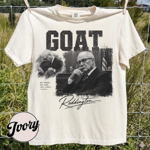 May include: An ivory short-sleeve t-shirt featuring a black and white graphic print. The design includes the word GOAT in bold letters, two images of a man in a suit, and the text: NOT POWER, NOT FAME, JUST LEGACY, and the signature Reddington.