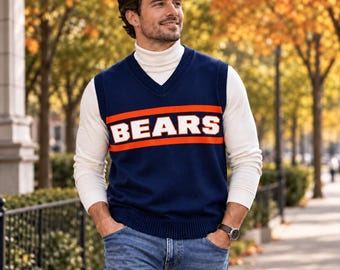 Op de afbeelding: Een marineblauw V-hals gebreid vest met het woord BEARS in grote witte blokletters tussen twee horizontale oranje strepen, gedragen over een wit shirt met lange mouwen en een col, gecombineerd met een blauwe spijkerbroek.