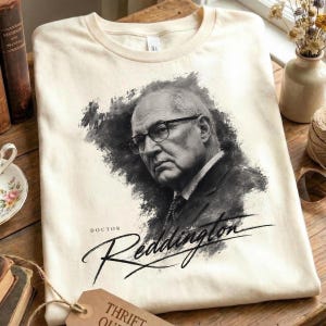 May include: A cream-colored t-shirt featuring a black and white artistic portrait of a man wearing glasses and a suit. Below the portrait, the text reads "DOCTOR" in small print and "Reddington" in a large, stylized, cursive script font.