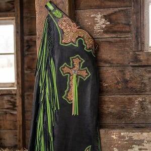 May include: Black leather chaps featuring long lime green and black fringe along the side. The chaps are decorated with tan floral tooling and a prominent cross emblem on the thigh, accented with lime green stitching and a central crystal detail.