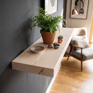 May include: A light-colored wooden floating shelf mounted on a dark gray wall. The shelf holds a green fern in a terracotta pot, a small succulent in a copper-colored pot, three smooth gray stones, and a small decorative bowl with a blue pattern.