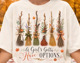 Puede incluir: Una camiseta color crema con un gráfico de seis escobas decorativas adornadas con follaje otoñal, calabazas y cintas. Debajo de las escobas, el texto dice, A Girl's Gotta Have OPTIONS, rodeado de pequeñas hojas de arce naranjas y una calabaza.