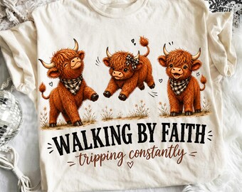 Puede incluir: Una camiseta color crema con un gráfico de tres vacas Highland marrones. Dos vacas llevan pañuelos a cuadros blancos y negros, y una tiene un lazo a juego. El texto dice "WALKING BY FAITH" en negro y "tripping constantly" en color marrón.