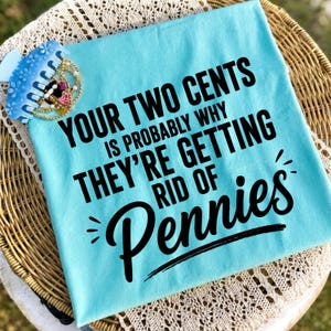 Op de afbeelding: Een lichtblauw t-shirt met zwarte tekst "YOUR TWO CENTS IS PROBABLY WHY THEY'RE GETTING RID OF Pennies" en een lichtblauwe haarklem versierd met kleine witte bloemetjes en goudkleurige kraaltjes op de stof.