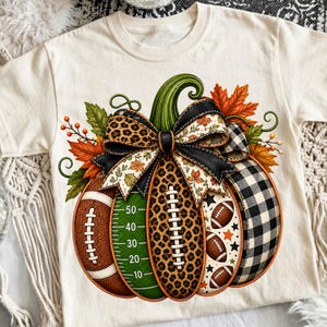 Pode incluir: Uma t-shirt de cor creme com um grande gráfico de uma abóbora decorada com motivos de futebol americano. Os segmentos da abóbora incluem uma textura de bola, um campo verde com linhas, estampado de leopardo e um padrão xadrez.