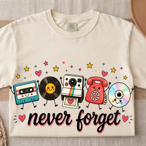 Puede incluir: Una camiseta de color crema con un gráfico de un casete, un disco de vinilo, una cámara instantánea, un teléfono de disco y un CD sonrientes. El texto "never forget" está impreso en negro debajo de los personajes, rodeado de estrellas.