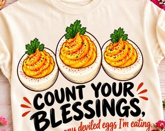 Puede incluir: Una camiseta de color crema con un gráfico de tres huevos rellenos decorados con perejil y pimentón. Debajo de los huevos, el texto negro dice "COUNT YOUR BLESSINGS," seguido del texto rojo "not how many deviled eggs I'm eating."