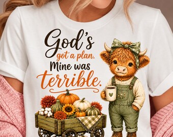 Puede incluir: Una camiseta blanca con el gráfico de una vaca highland esponjosa con peto verde y un lazo, sosteniendo una taza. Debajo, un carro de madera lleno de calabazas, girasoles y flores. El texto dice: God's got a plan. Mine was terrible.