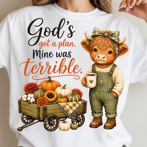May include: A white t-shirt featuring a graphic of a highland cow wearing green overalls and a bow, holding a mug. The text reads, God's got a plan. Mine was terrible. Below is a wooden wagon filled with orange and green pumpkins and flowers.