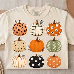 May include: A cream-colored t-shirt featuring a grid of nine pumpkins with various patterns including leopard print, black and white plaid, white flowers on orange, black polka dots, solid orange, green with vines, orange stripes, black with white flowers, and orange with white hearts.