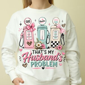 May include: A white long-sleeve sweatshirt featuring a graphic of three vintage gas pumps in pink, teal, and cream. The design includes floral accents, leopard print hearts, and bows, with the text "THAT'S MY Husband's PROBLEM" printed in pink and teal.
