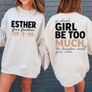 May include: A cream-colored crewneck sweatshirt featuring black and tan text. The front displays ESTHER four fourteen with bows and a cross. The back displays go ahead, GIRL BE TOO MUCH, and the kingdom needs your voice in black and tan.