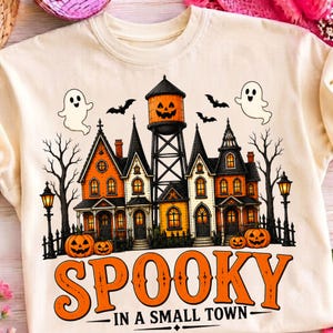 May include: A cream-colored t-shirt featuring a graphic of a haunted house with orange windows, a water tower with a jack-o'-lantern face, two ghosts, bats, and pumpkins. The text below reads SPOOKY IN A SMALL TOWN in large orange letters.
