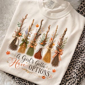 May include: A cream-colored t-shirt featuring a graphic of six decorative brooms adorned with autumn foliage, pumpkins, and ribbons. Below the brooms, the text reads, A Girl's Gotta Have OPTIONS, surrounded by small orange maple leaves and vines.