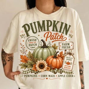 Pode incluir: Uma t-shirt cor de creme com uma impressão gráfica com o texto PUMPKIN Patch, FRESH FROM THE PATCH, FARM FRESH, LOCALLY GROWN, AUTUMN HARVEST, PUMPKINS, CORN MAZE e APPLE CIDER, juntamente com ilustrações de abóboras, folhas e um girassol.