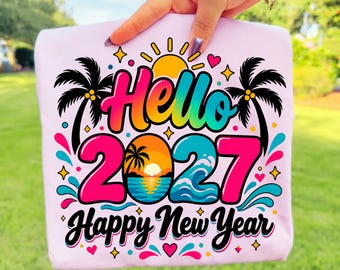 Puede incluir: Una camiseta rosa claro con un gráfico colorido que incluye el texto Hello 2027 Happy New Year. El diseño presenta un sol amarillo, siluetas de palmeras negras, olas del mar y pequeños corazones y estrellas en tonos vibrantes de rosa, azul y naranja.